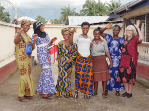 Teilnehmerinnen eines Alphabetisierungskurses in einem Schulhof in Sierra Leone, in bunten Kleidern teils mit traditionellen und/oder muslimischen Kopftüchern, mit Koordinatorin Sigrid Schell-Straub